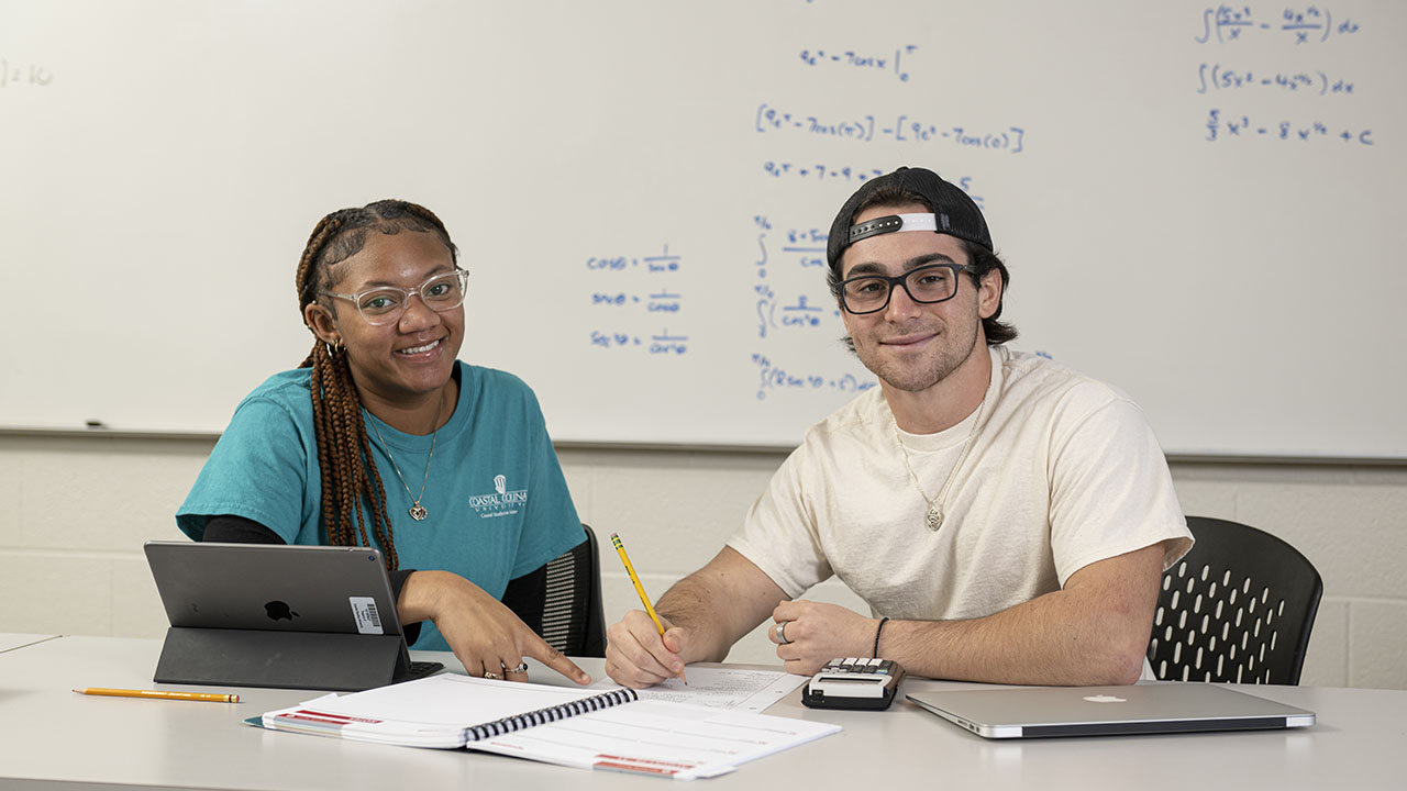 Two students sit at a table working together on math problems, using notebooks, a calculator, and a tablet in a classroom setting with equations written on a whiteboard behind them.